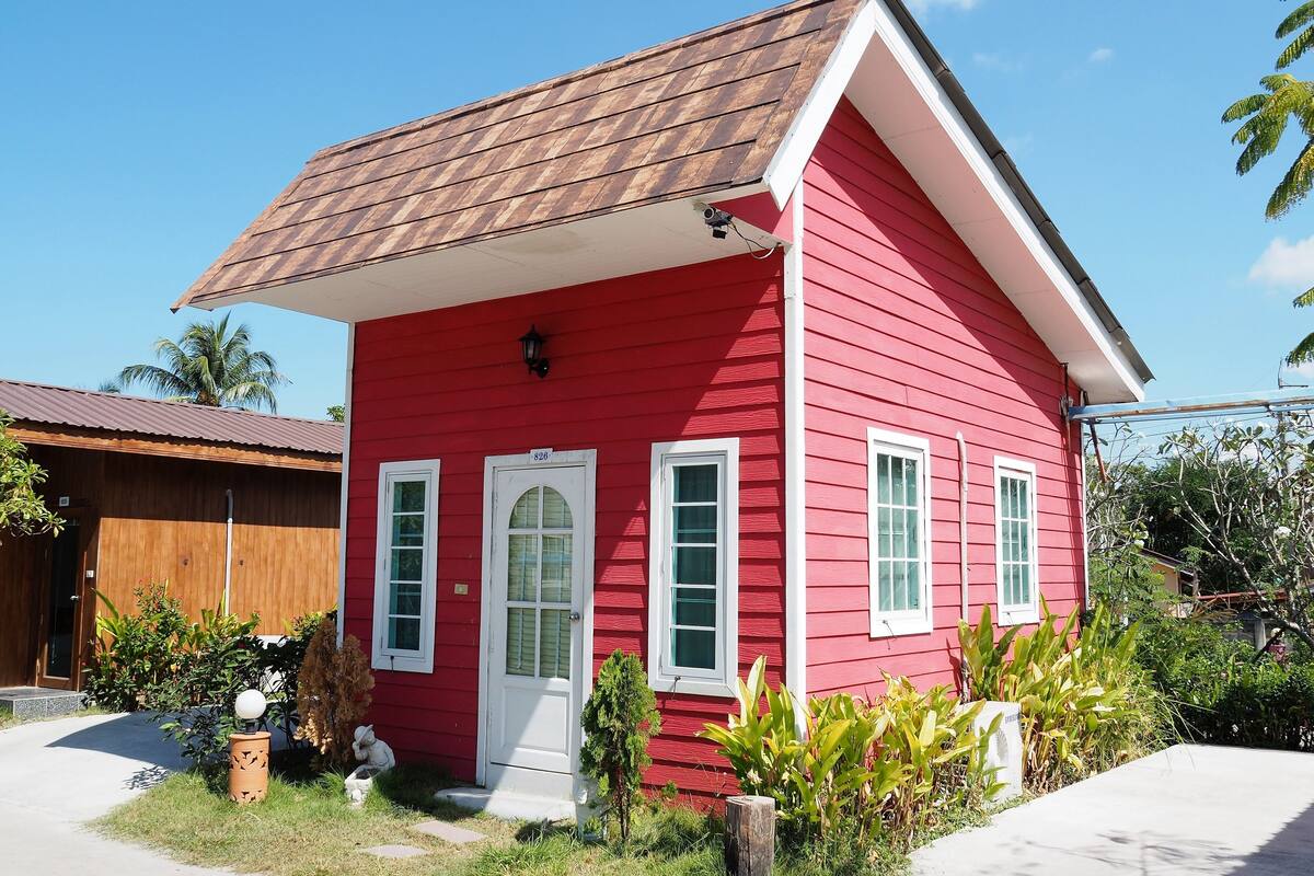 A red house with a brown roof, white door, and windows, surrounded by greenery.