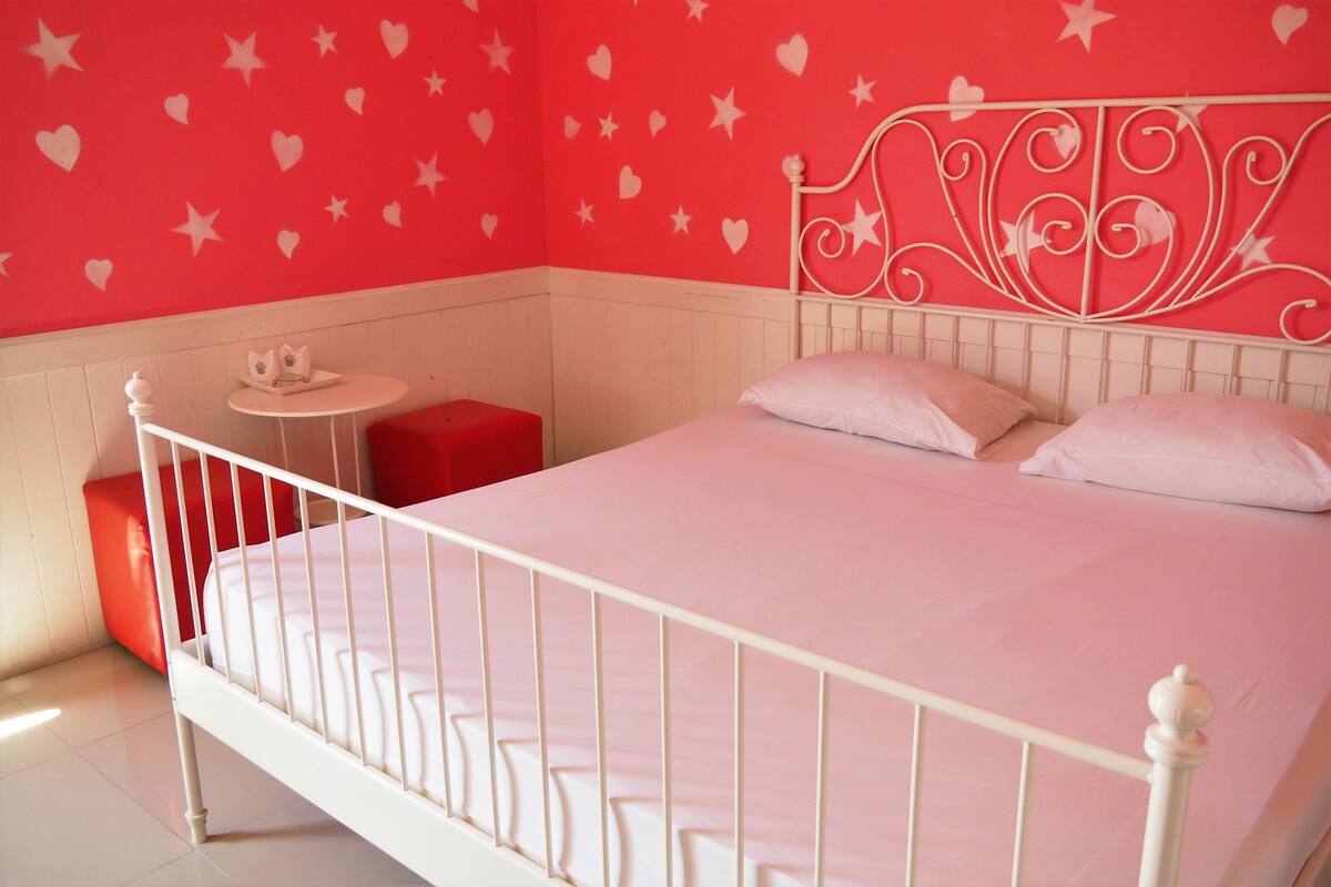 A white metal bed with a headboard, a red bench, and a small round table with a white object on it.