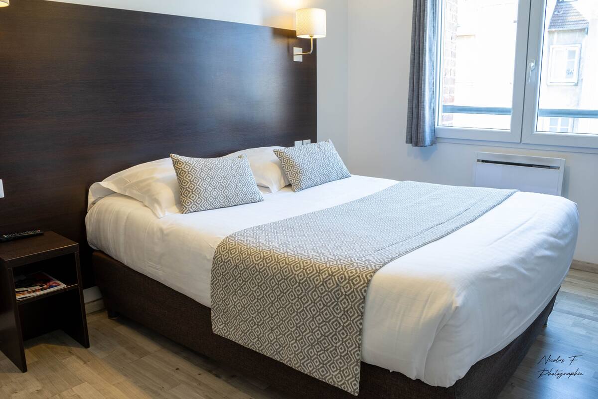 Una cama bien hecha con almohadas estampadas y una colcha, una mesita de noche con una revista, y una ventana con cortinas.