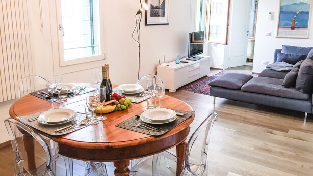 A dining area with a wooden table set for four, surrounded by clear chairs. A television and a sofa are visible in the background. - Cà Farnese (Venice)