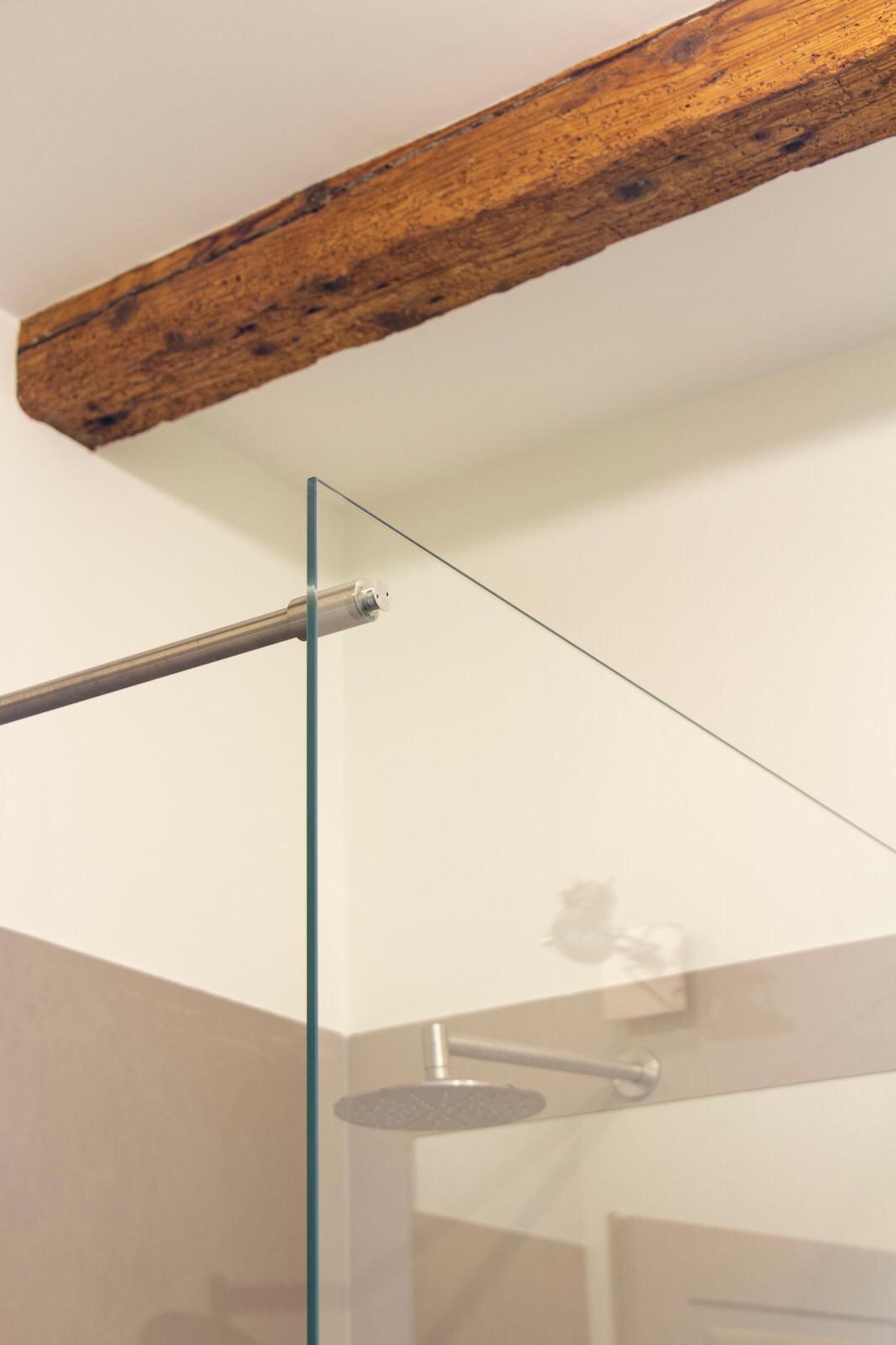 A glass shower enclosure with a wooden beam above, a metal handle, and a visible showerhead.