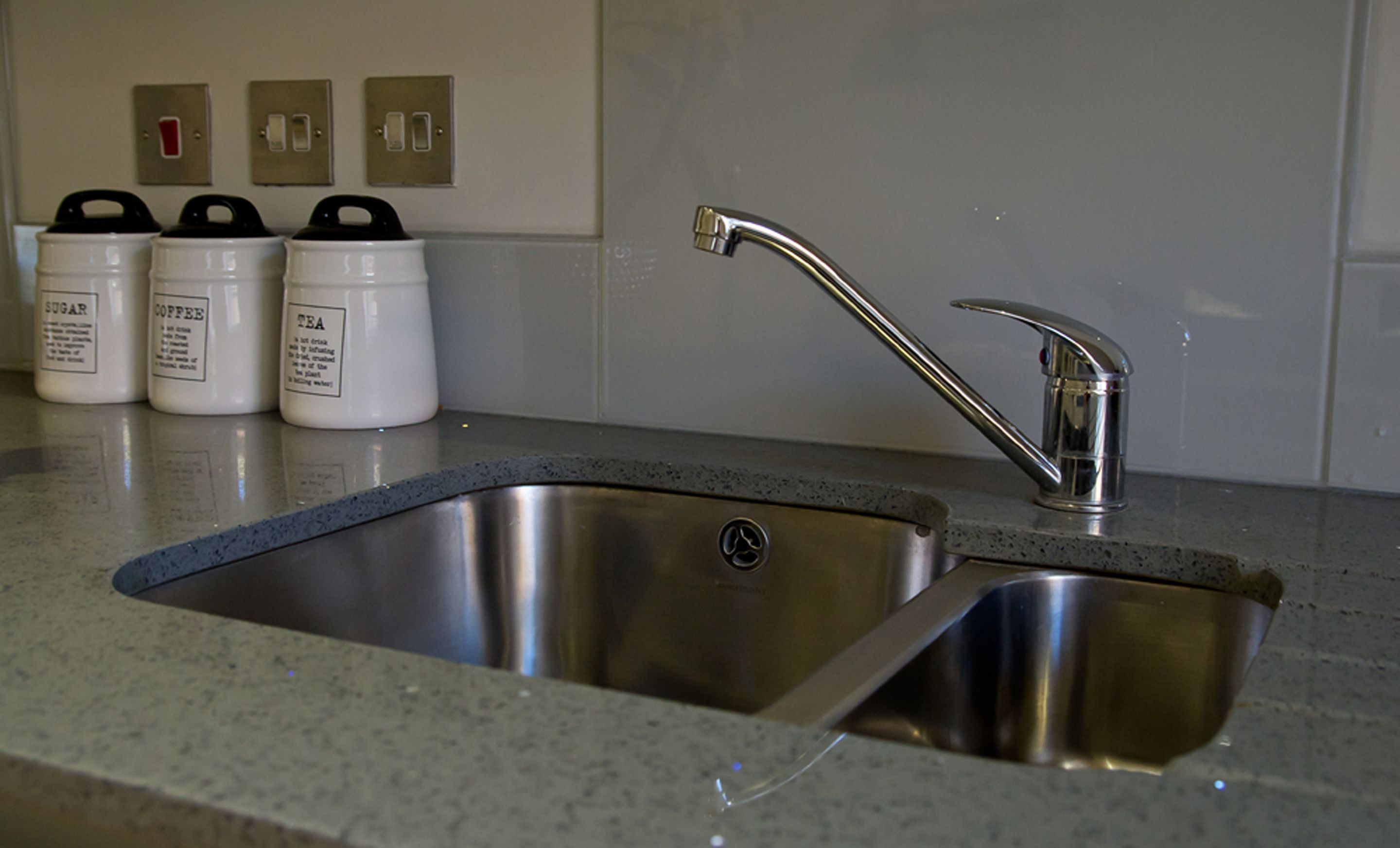 A stainless steel kitchen sink with a modern faucet, three white containers labeled sugar, coffee, and tea, and three wall switches above the containers.