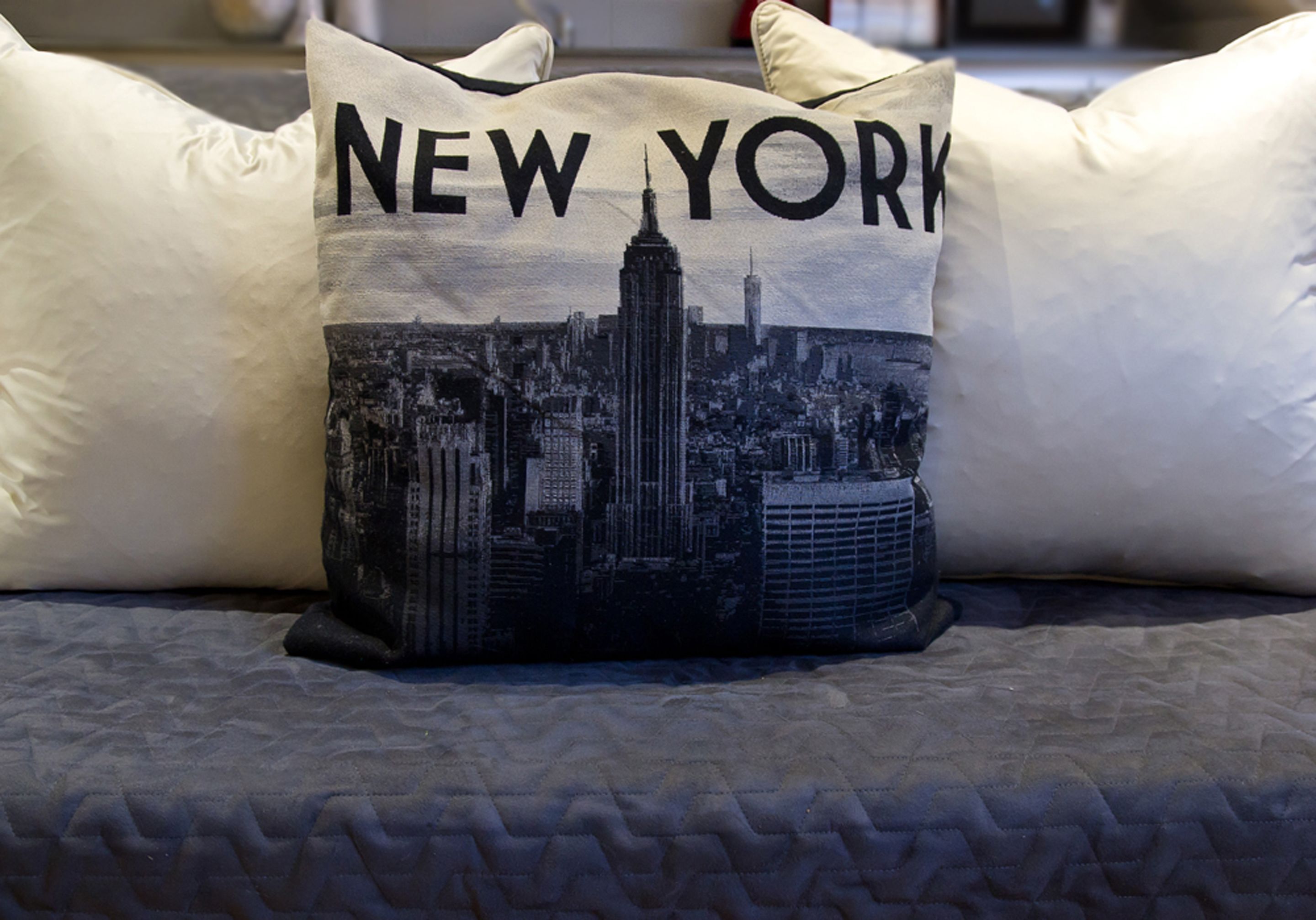 A pillow with a New York City skyline print, placed on a bed with a quilted cover.
