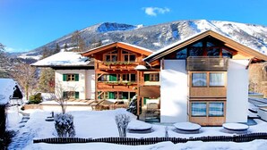 A modern chalet with a wooden facade, large windows, and a balcony overlooking a snowy mountain landscape.