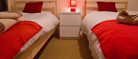 A twin room with two beds, each with a red blanket and pillow, a bedside table with a lamp, and a wooden headboard.