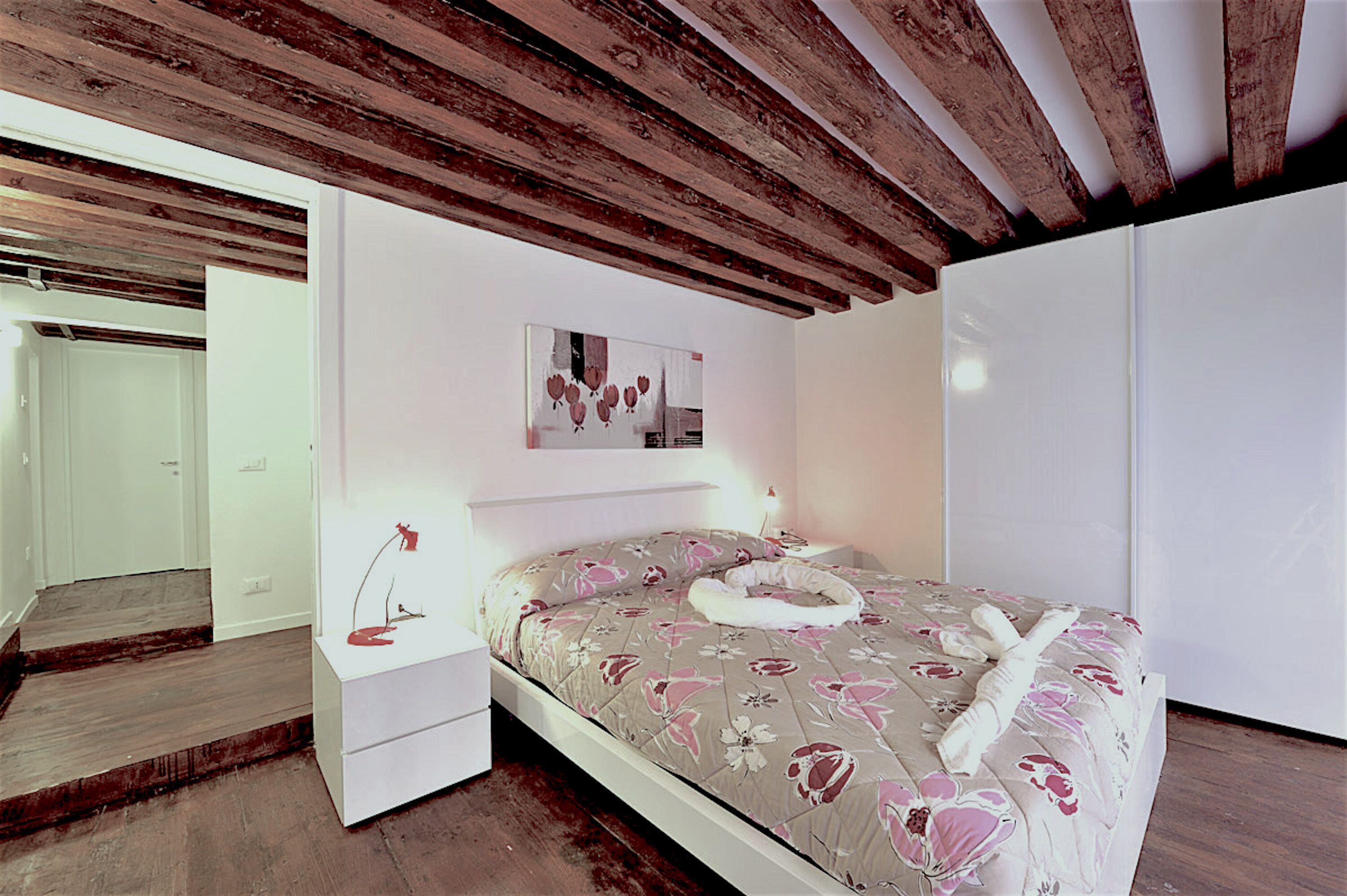 Une chambre avec un plafond en bois, un lit recouvert d’une literie à motifs floraux, une table de chevet avec une lampe et un tableau accroché au mur.
