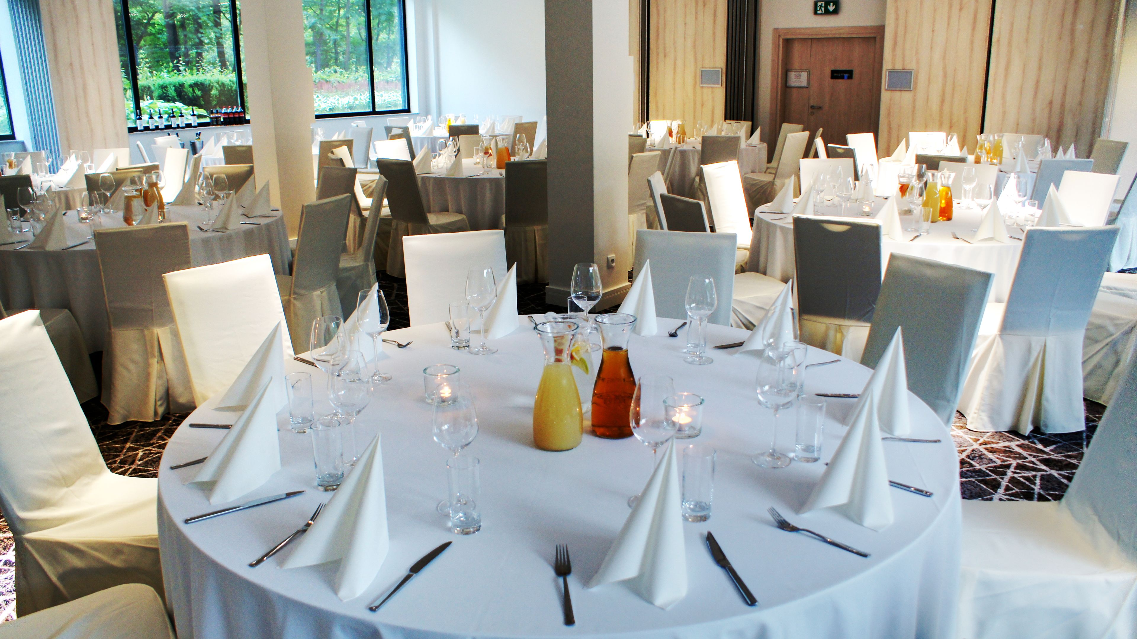 A formal dining room with round tables set for a meal, each with white tablecloths, glassware, and silverware.