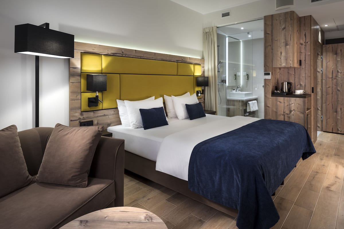 Ein modernes Hotelzimmer mit einem großen Bett, einer Couch, einem Nachttisch mit Lampe, einem Holzkopfteil und einem durch eine Glastür einsehbaren Badezimmer.