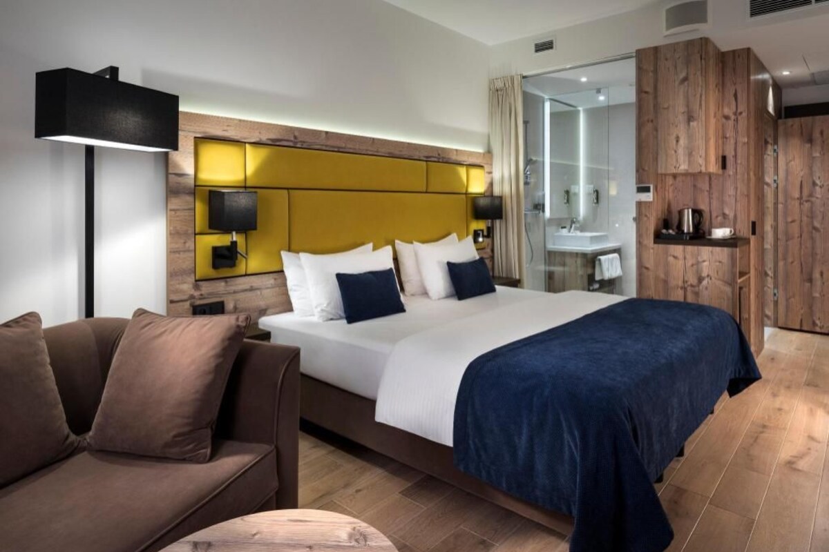 A modern hotel room with a large bed, a sofa, a bedside table, a lamp, and a wooden wardrobe.