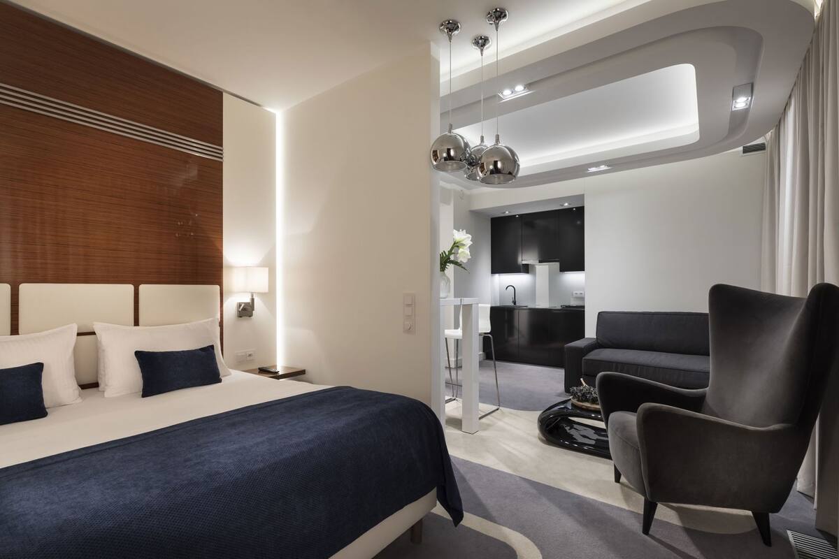 Ein modernes Hotelzimmer mit einem großen Bett, einem dunklen Sessel, einem kleinen Tisch und einer Küchenzeile.