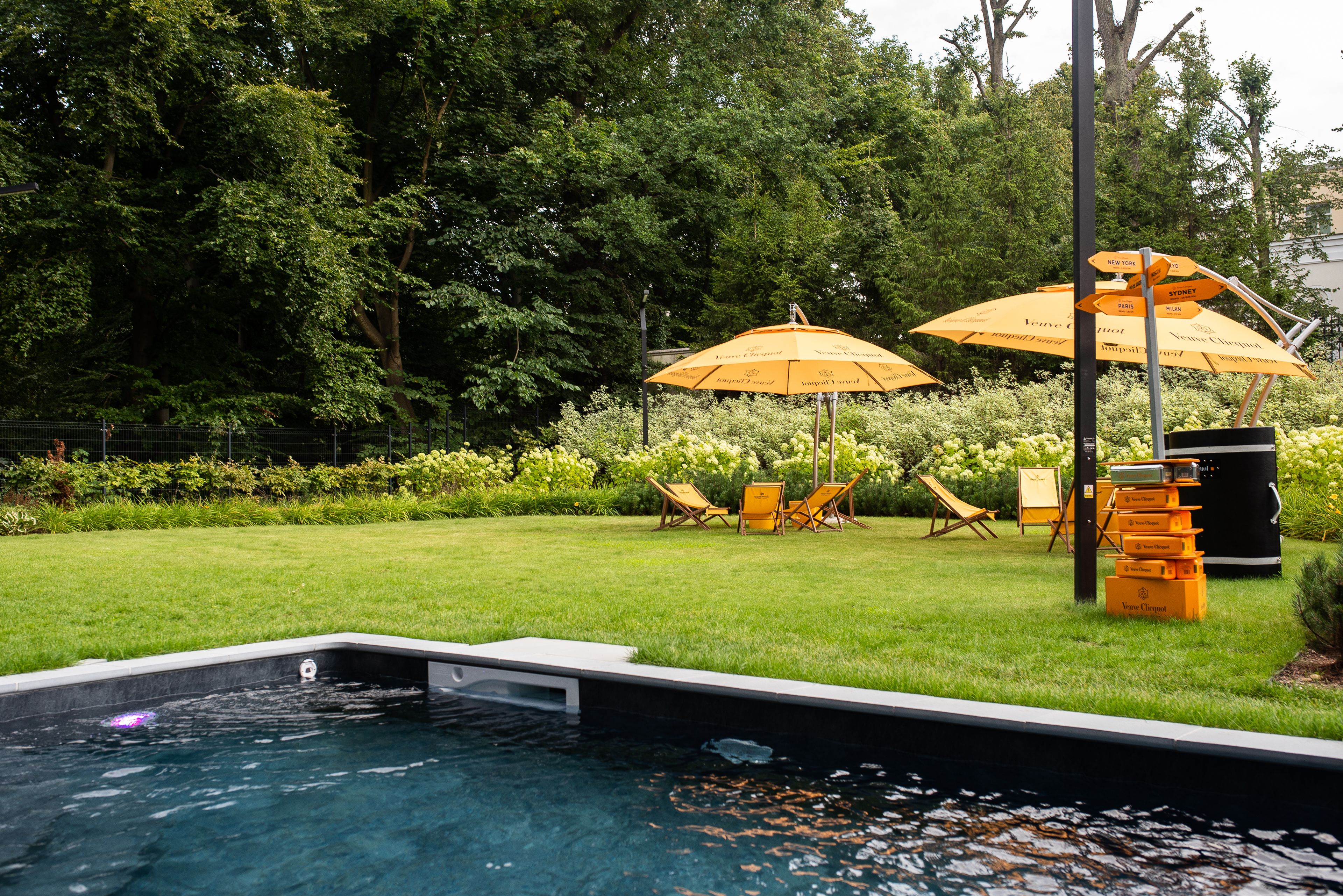 A swimming pool with a small deck, surrounded by grass and trees. There are two yellow umbrellas and lounge chairs.
