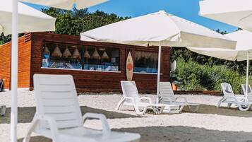 A beachside kiosk with white plastic chairs and umbrellas, a wooden structure with a surfboard sign, and a sandy beach with greenery in the background.