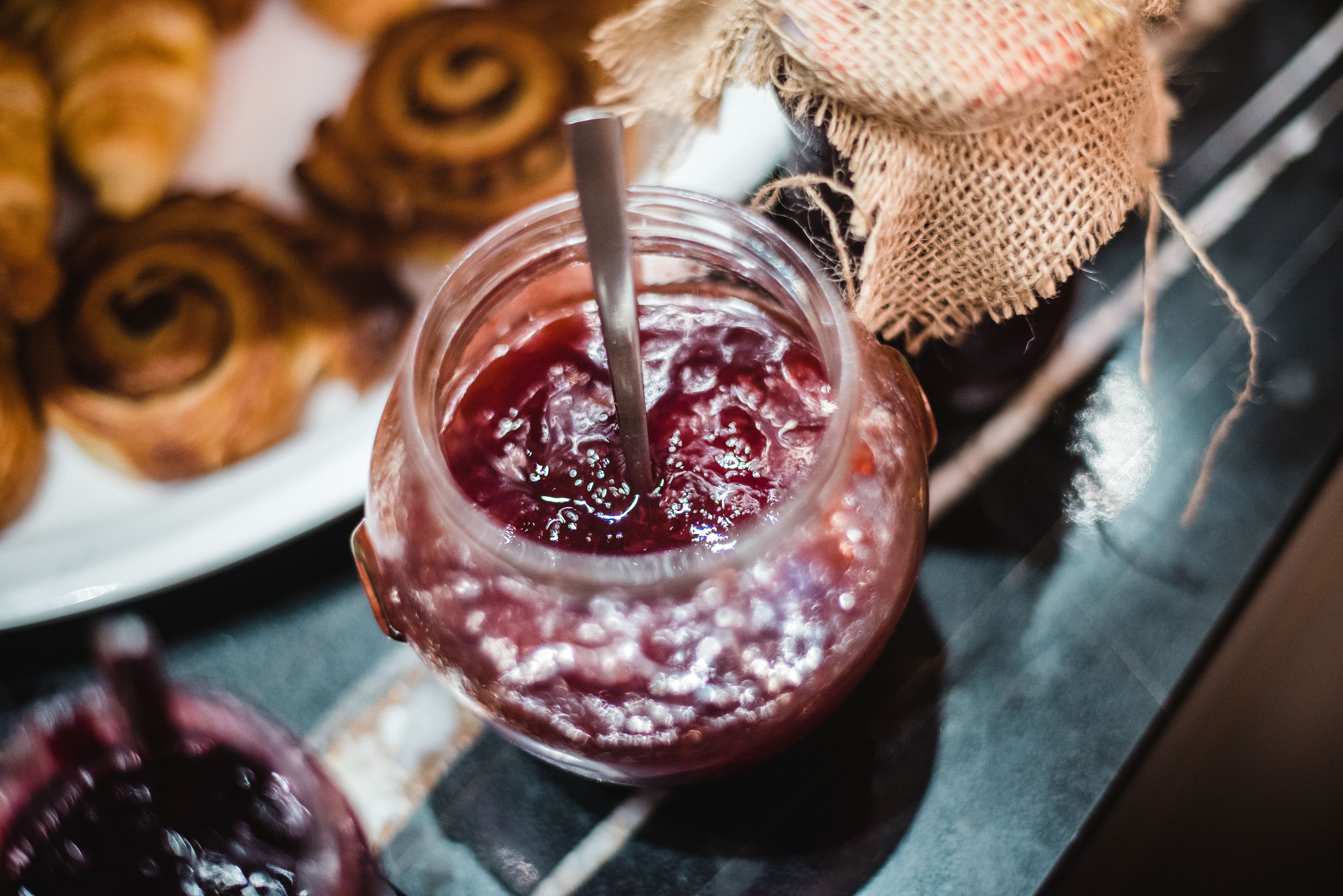 A jar of jam with a spoon, a burlap sack, and a plate of pastries.
