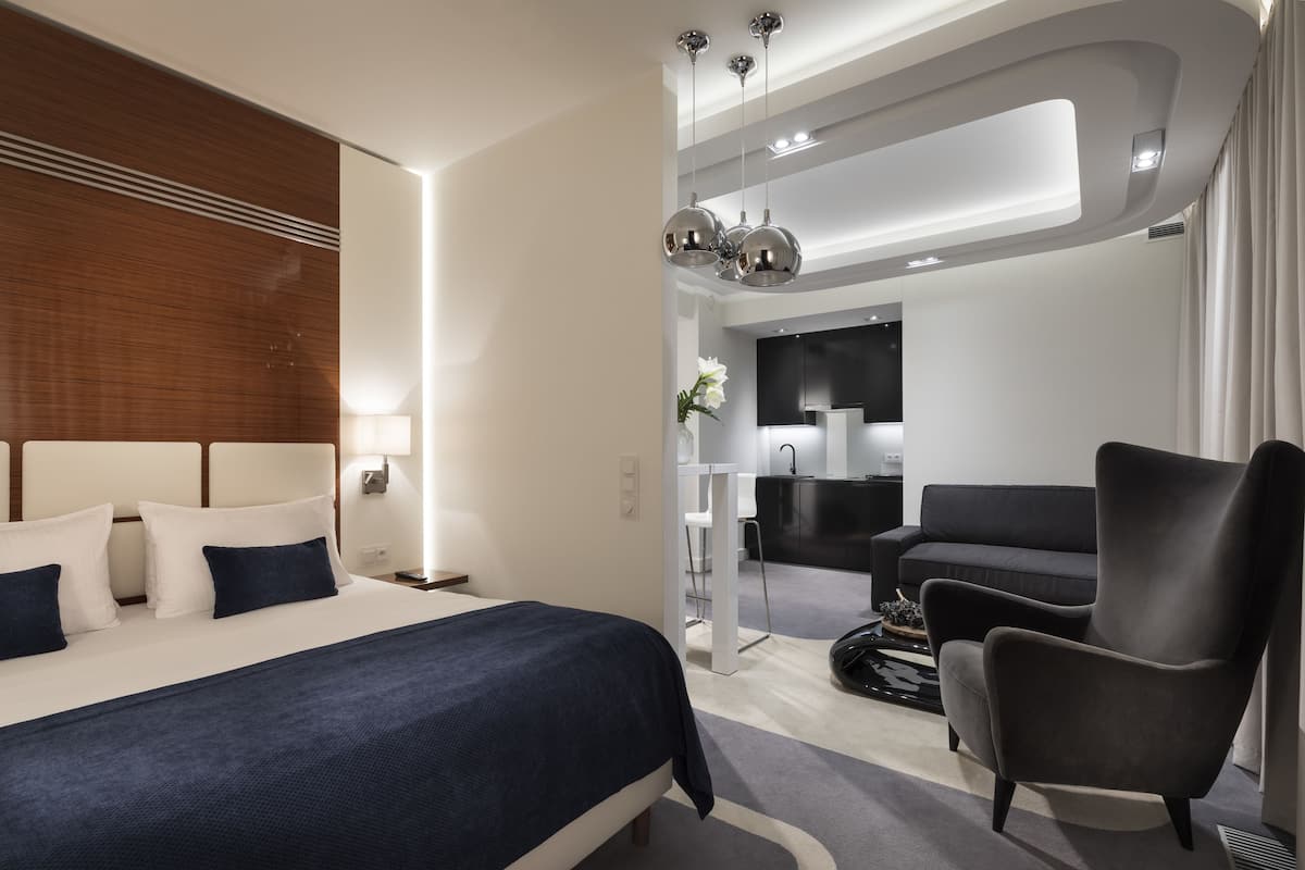 Ein modernes Hotelzimmer mit einem großen Bett, einer Sitzecke mit einem Sessel, einem Schreibtisch und einer Küchenzeile.