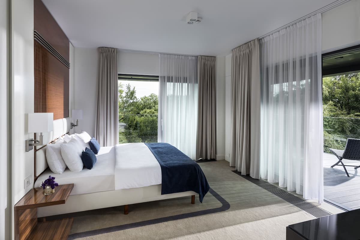 Ein modernes Hotelzimmer mit einem großen Bett, einem Nachttisch mit Lampe und Blick ins Freie durch große Fenster.