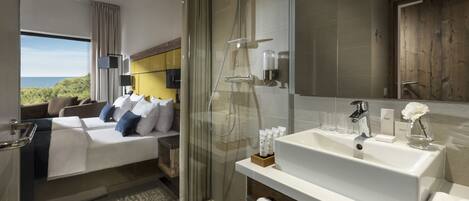 A modern hotel room with a large bed, a glass-enclosed shower, a white sink, and a view of the outdoors.