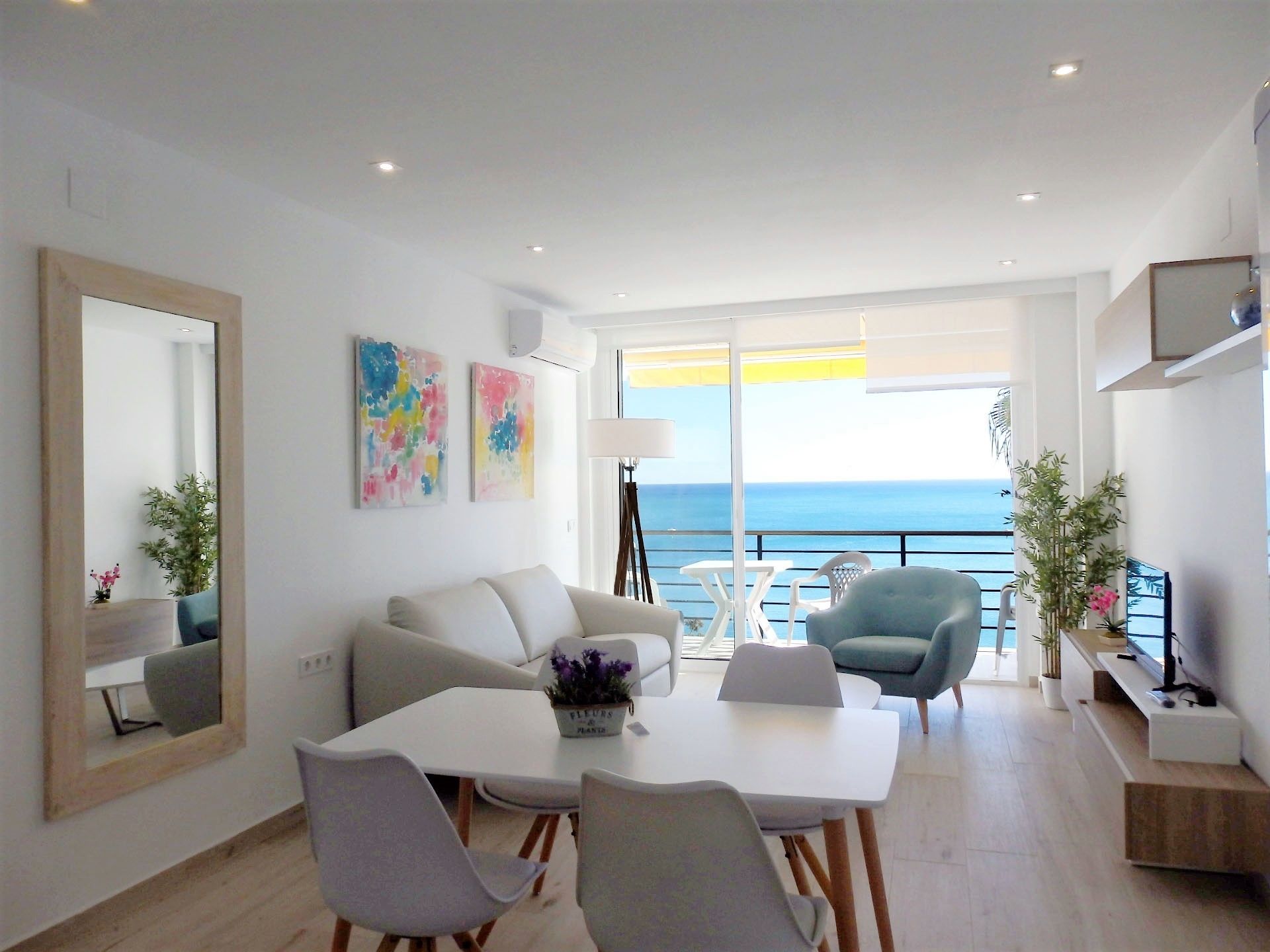 Een moderne woonkamer met een balkon en uitzicht op zee, een eethoek met een tafel en stoelen, en een televisie op een standaard.