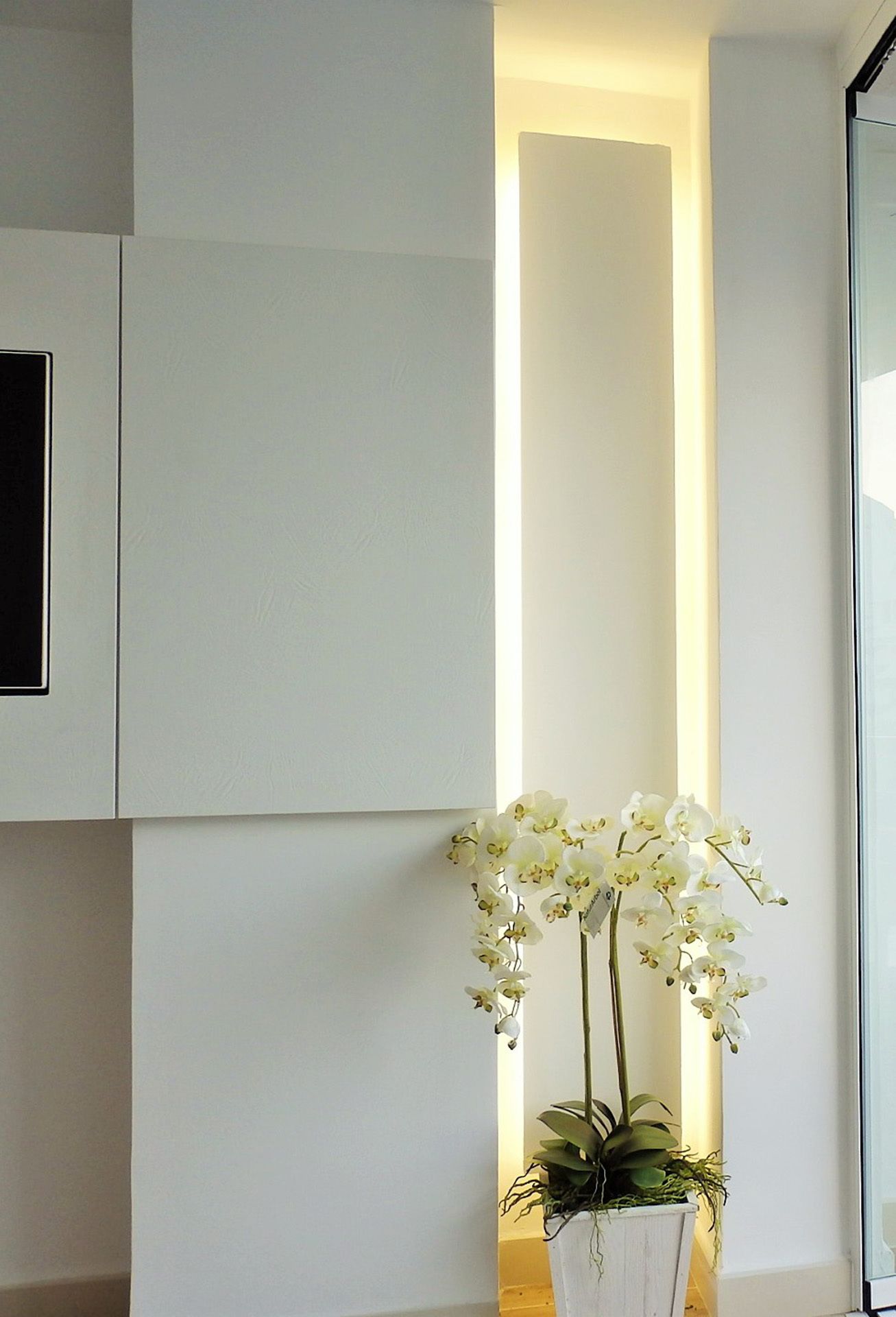 A modern interior with a vertical light panel, a white orchid plant, and a white cabinet.