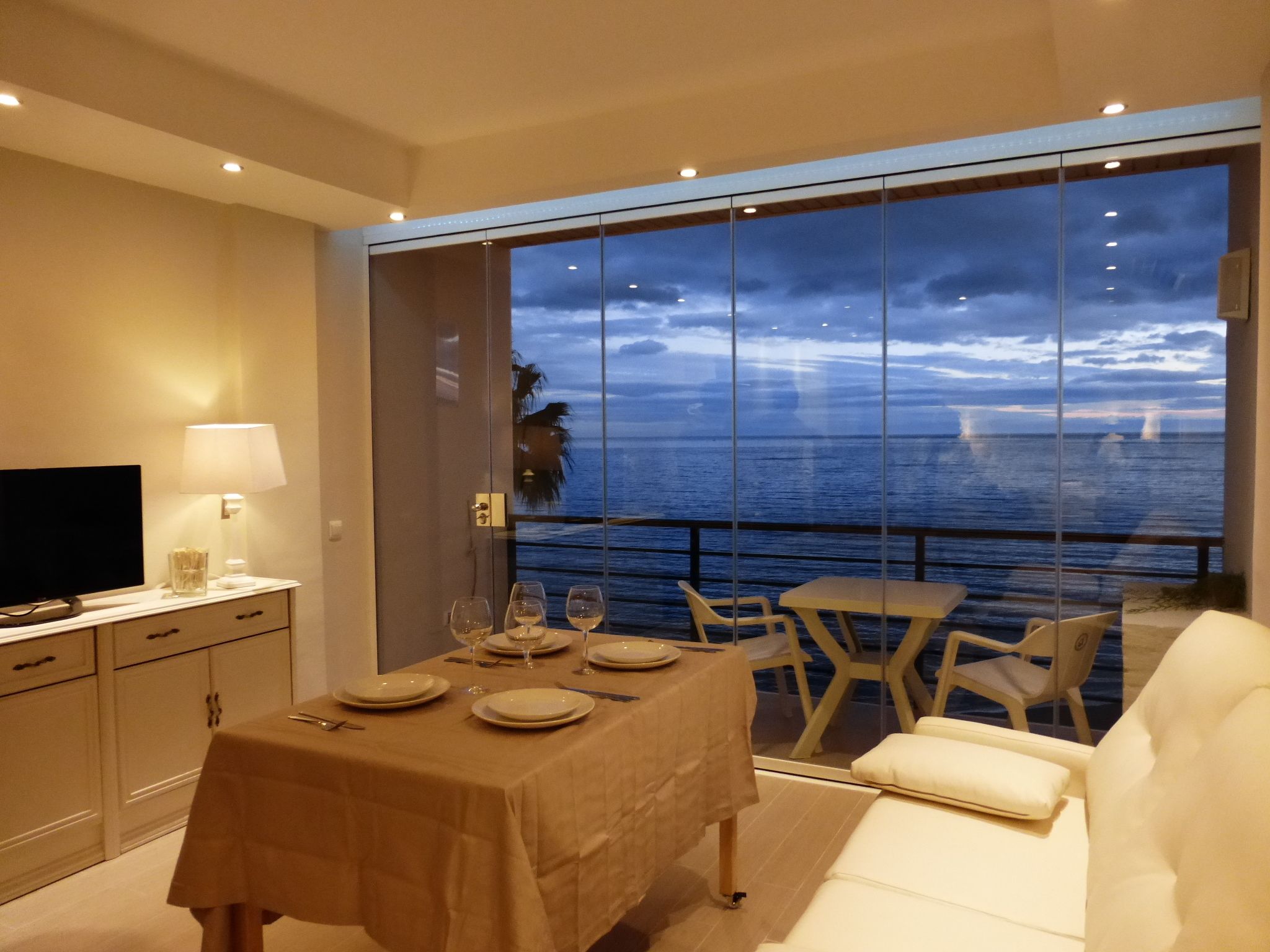A dining area with a table set for two, a television, and a view of the ocean through large windows.