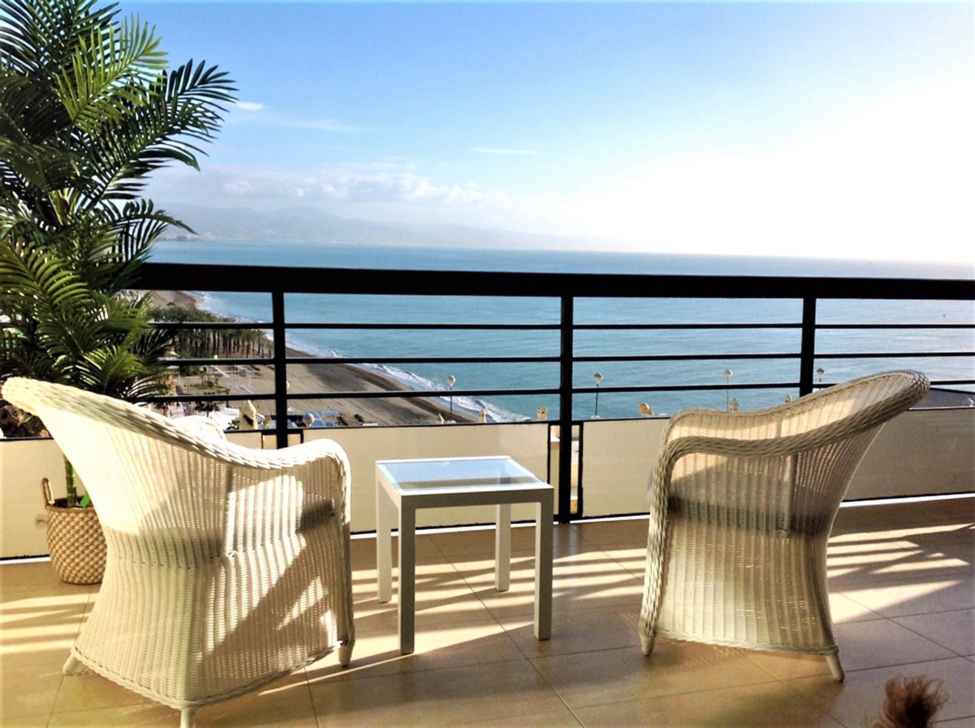 A balcony with wicker chairs and a small table, overlooking a beach and the ocean.