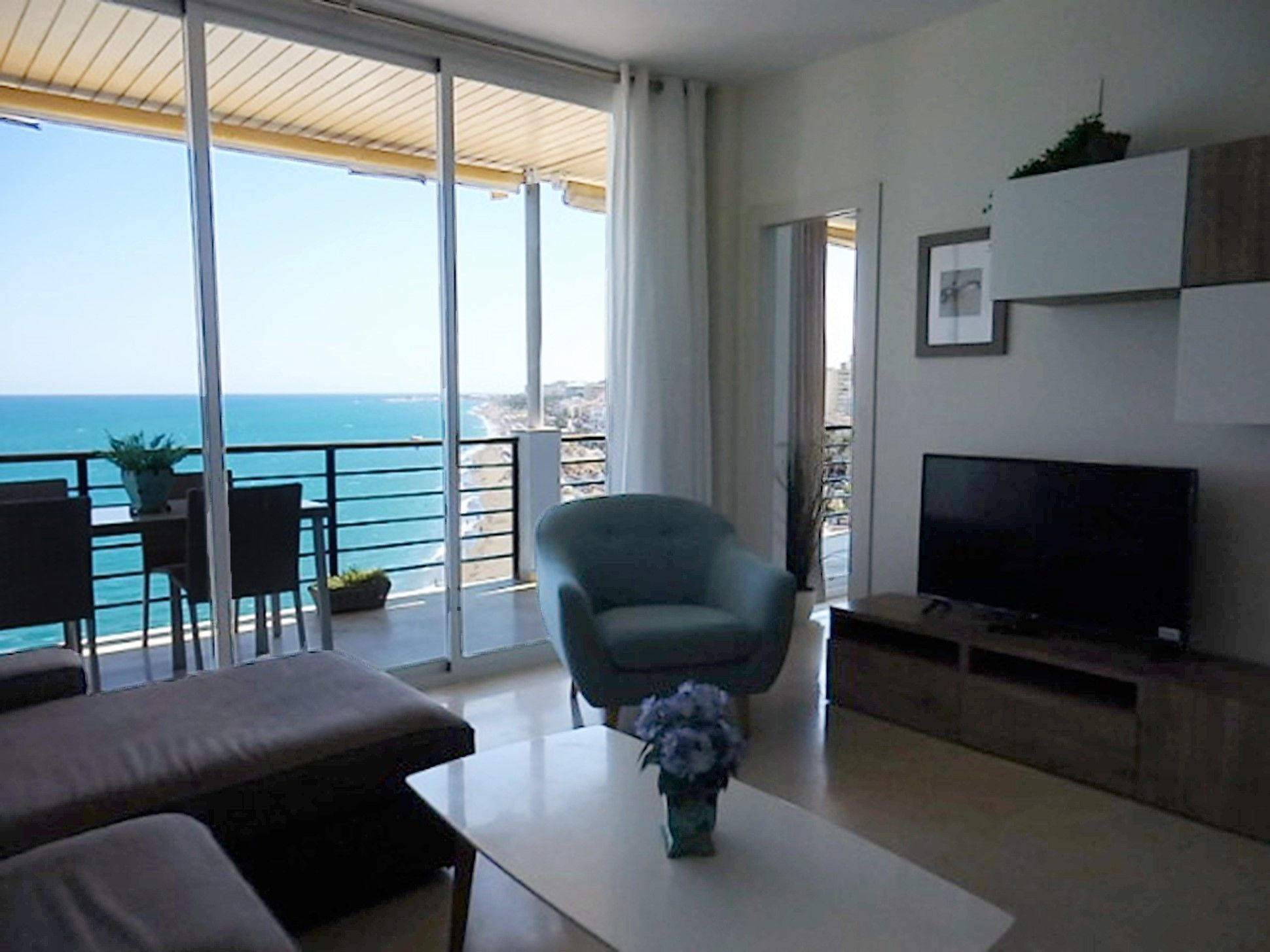 A modern living room with a large TV, a sofa, a chair, and a coffee table. There is a balcony with a view of the sea.