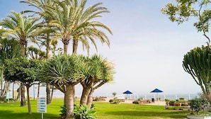 A landscaped garden with palm trees, a sign listing plant species, and a view of the ocean. - Waterfront Santa Clara Apartments (Torremolinos)