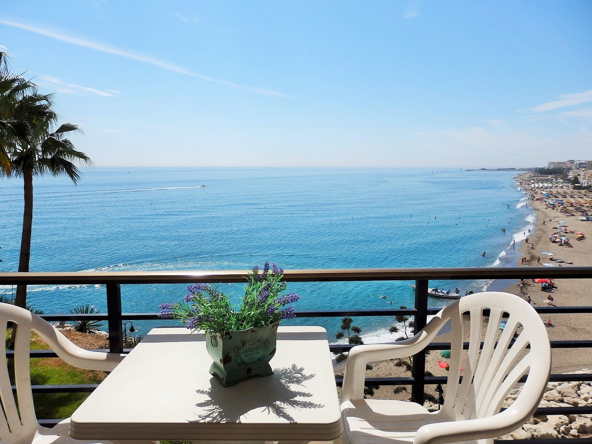 Een balkon met een tafel en stoelen, met uitzicht op een strand met mensen en palmbomen.