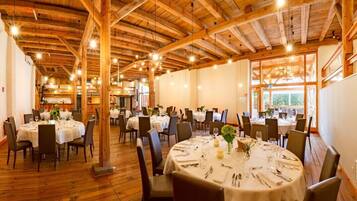 Een restaurant met ronde tafels klaar voor een maaltijd, houten balken aan het plafond en grote ramen voor natuurlijk licht.