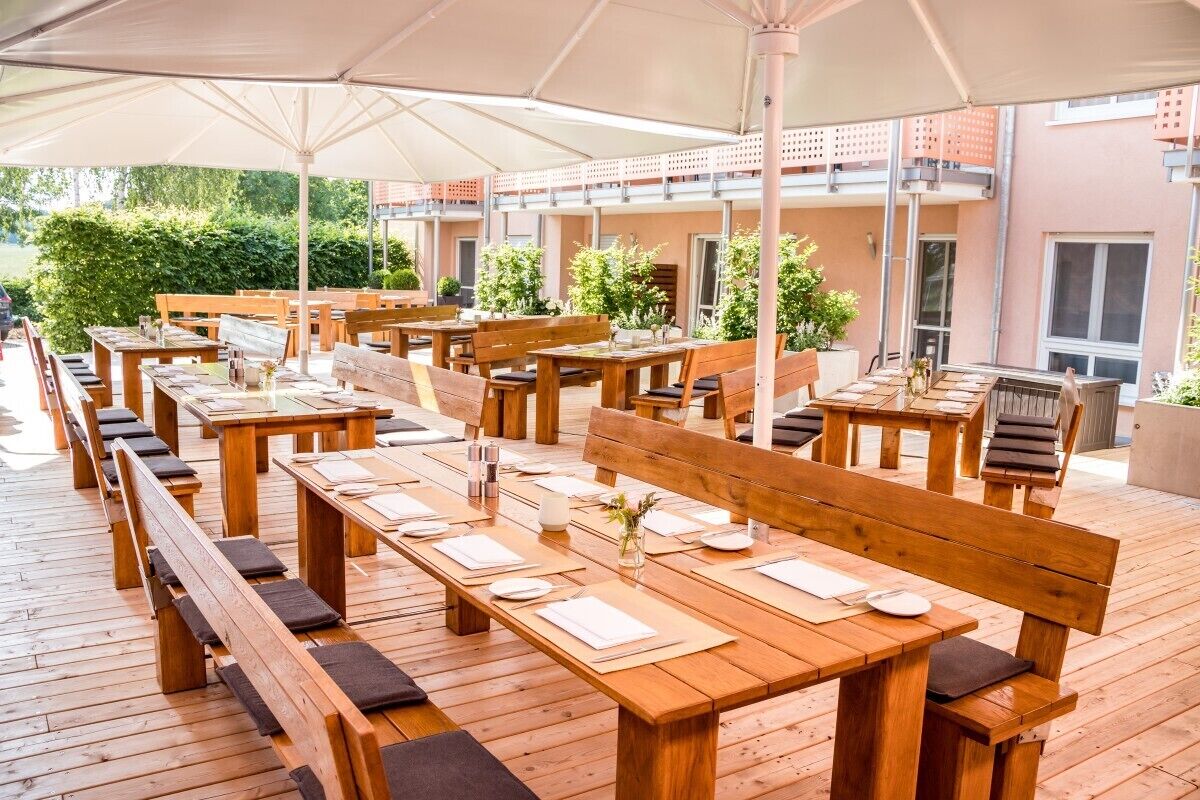 Een buiten eetgedeelte met houten tafels en banken, overdekt door grote parasols, en omgeven door een gebouw met een balkon.