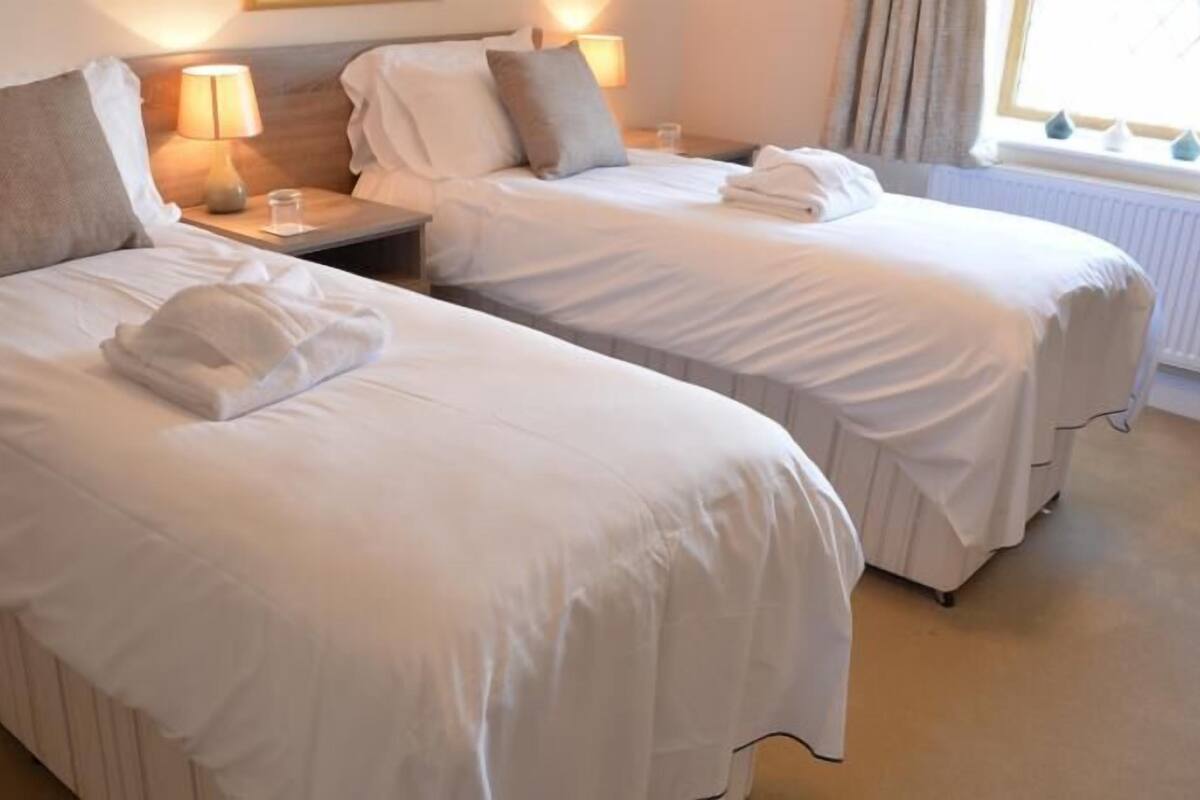 Ein Hotelzimmer mit zwei Einzelbetten, jeweils mit einem Kopfkissen und einem zusammengeklappten Handtuch. Ein Nachttisch mit einer Lampe und einem Glas Wasser ist ebenfalls vorhanden.