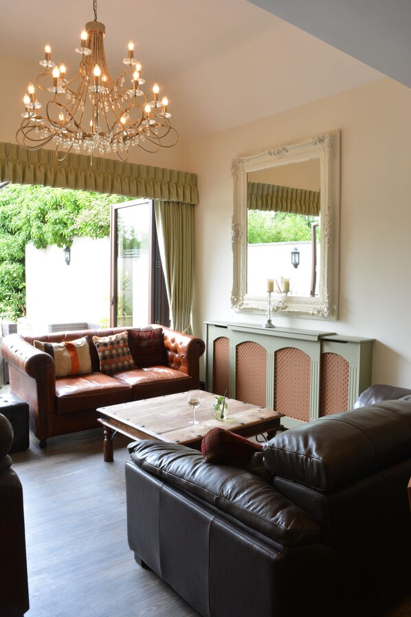 A living room with a chandelier, a brown leather sofa, a black leather couch, and a table with a glass of wine.