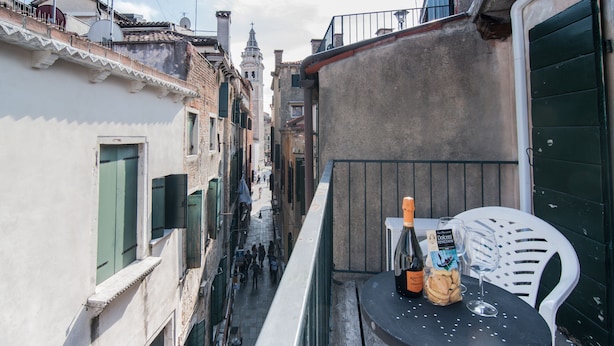 Une rue étroite bordée de bâtiments des deux côtés, un balcon avec une table et des chaises, et une bouteille de vin.