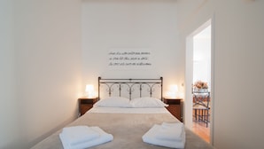 Un lit bien fait, avec du linge de lit et des serviettes blanches, une citation décorative accrochée au mur et, par une porte ouverte, une vue sur un coin repas.