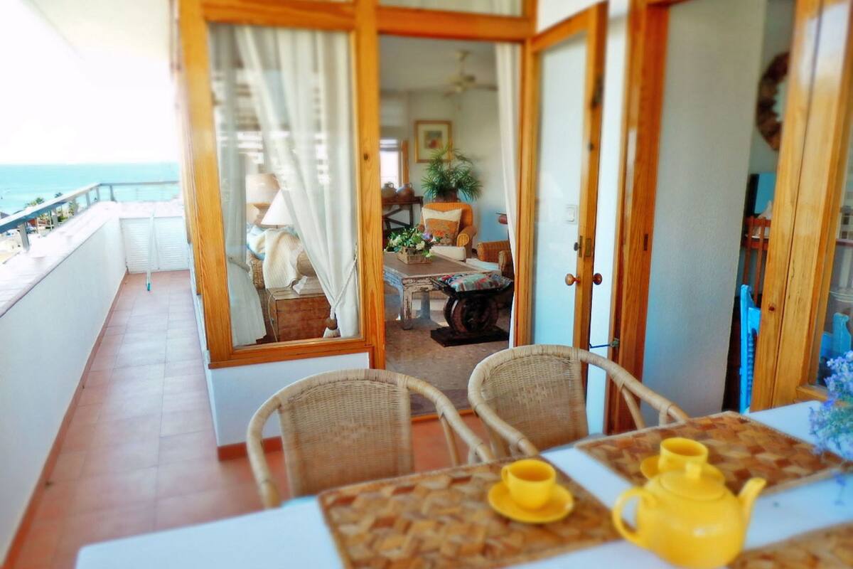 Balcó amb vista al mar, cadires de vimet, taula amb una tetera groga, i un interior acollidor amb un sofà i una planta.