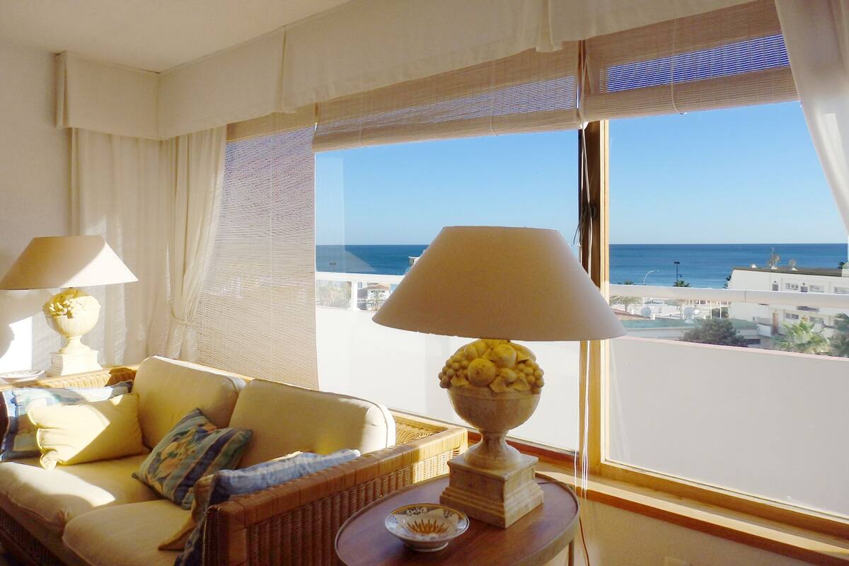 Habitació amb un sofà, dues làmpades, un plat de fruita i vista al mar.