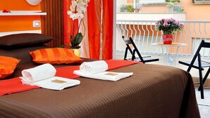 Ein Hotelzimmer mit einem großen Bett, orangefarbener Bettwäsche, einer Topfpflanze mit Orchideen und einem Balkon mit Stuhl und Tisch.