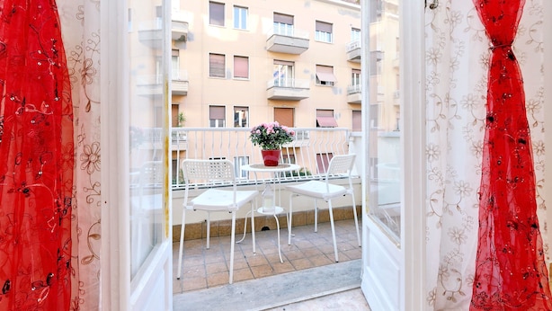 Ein offener Balkon mit Tisch und Stühlen, einem Blumentopf und Blick auf ein Gebäude mit Balkonen.