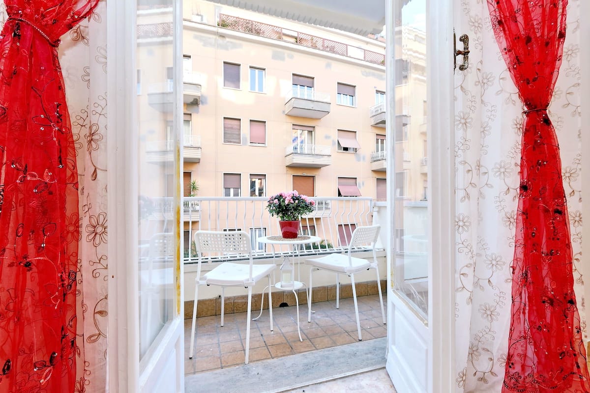 An open balcony with a table and chairs, a potted plant, and a view of a building with balconies.