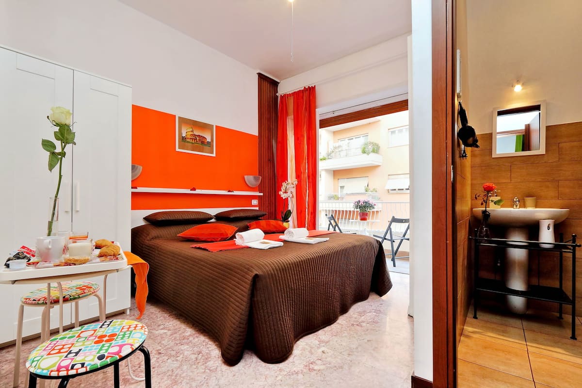 A hotel room with a bed, a desk with a chair, a small table, a bathroom with a sink, and a view of the outside balcony.