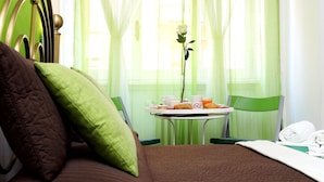 Ein Hotelzimmer mit einem Bett, einem grünen Sessel, einem Tisch mit Frühstücksgebäck und einem Fenster mit transparenten Vorhängen.