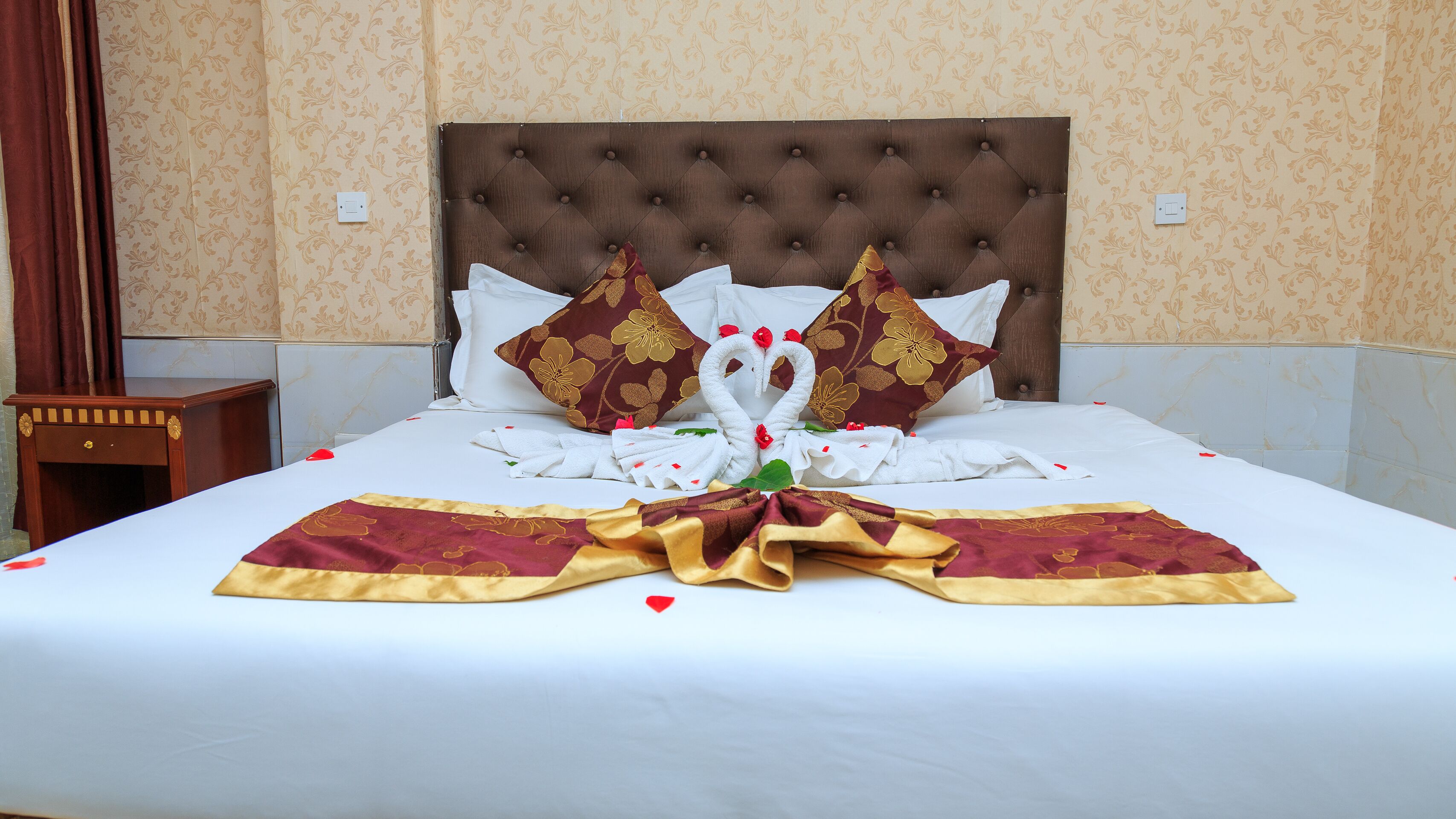 A neatly made bed with a decorative swan towel arrangement, a wooden nightstand, and patterned wallpaper.