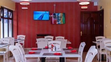 A dining area with white chairs and tables, red patterned wallpaper, and two paintings on the wall.