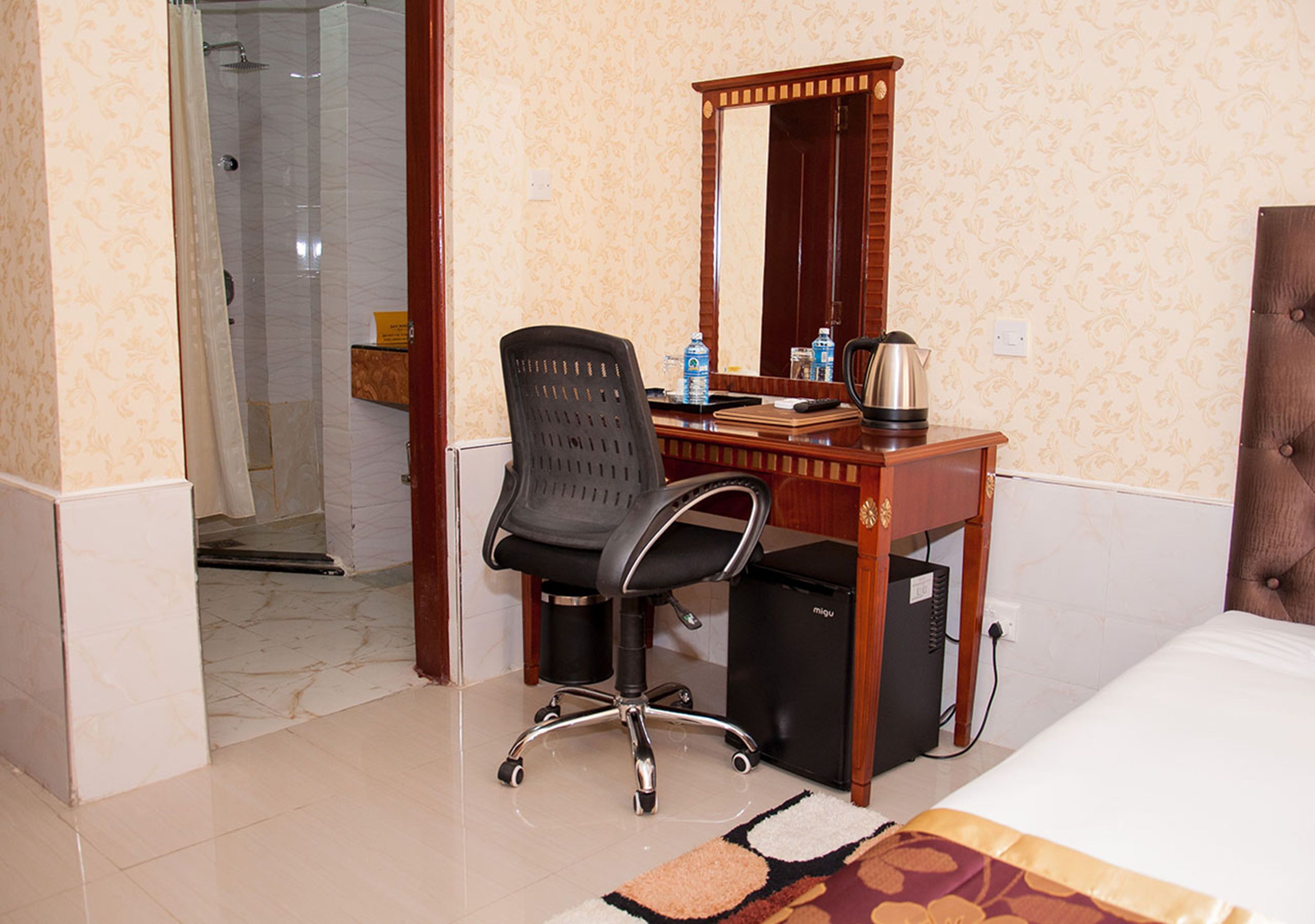 A hotel room with a bed, a desk with a chair, a mirror, a small refrigerator, and a shower area.