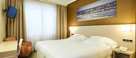 A hotel room with a large bed, a desk with a chair, a TV mounted on the wall, and a painting of a coastal scene.
