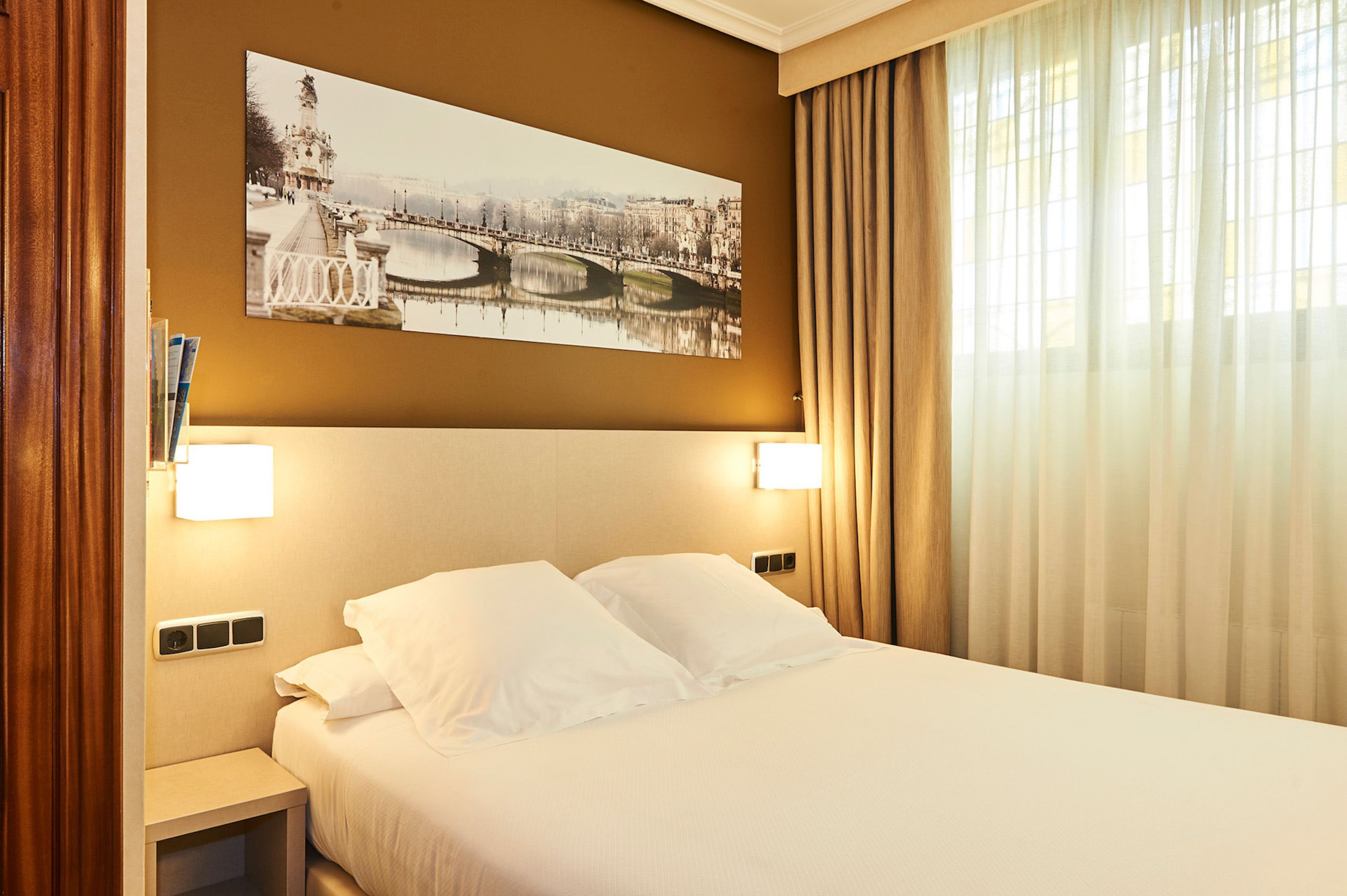 Una habitación de hotel con cama, mesita de noche, lámpara de pared y un amplio ventanal con cortinas.