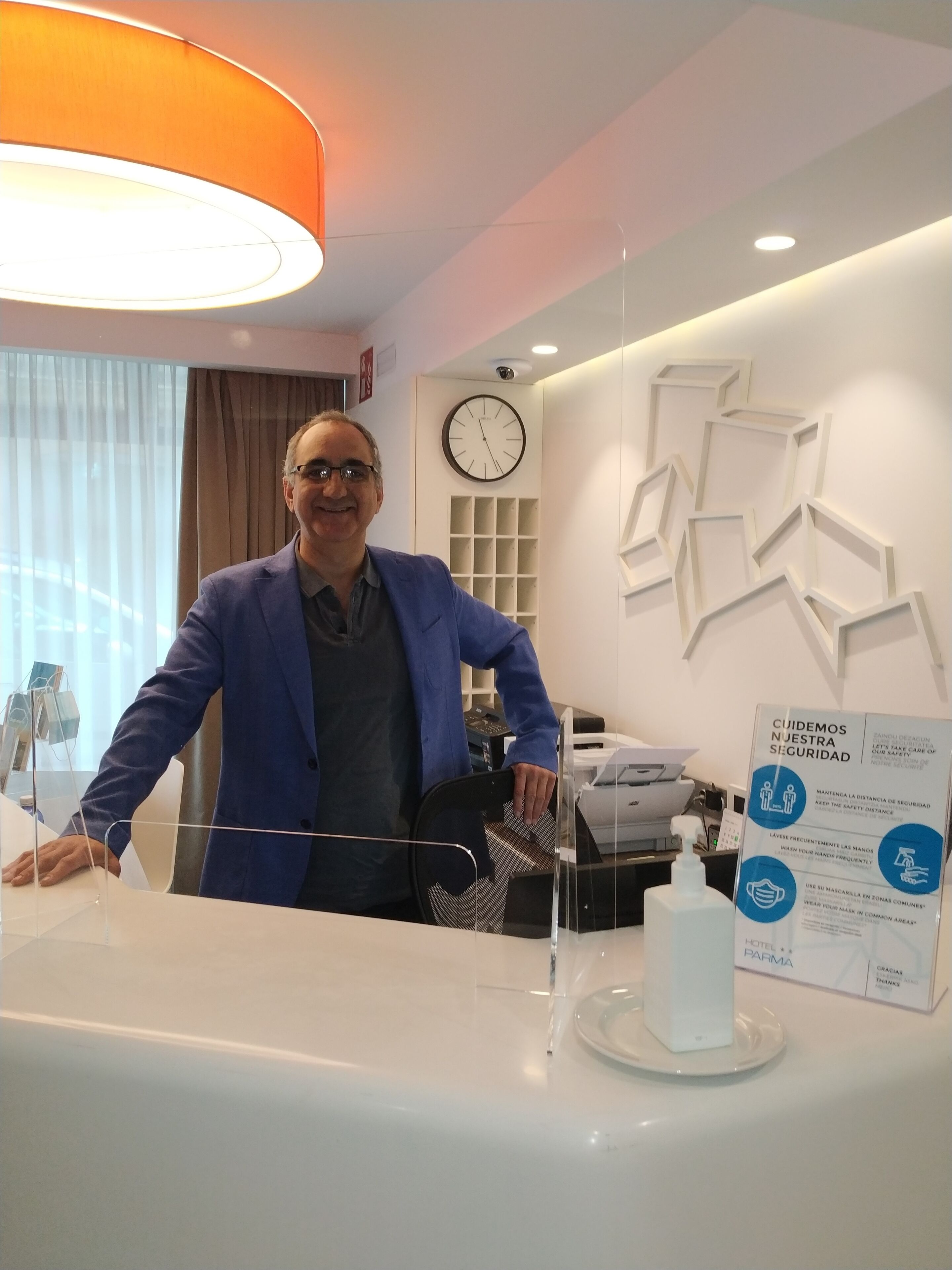 A hotel reception area with a modern design, including a reception desk, a clock, and a sign with health and safety guidelines.