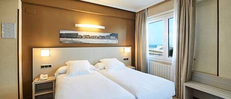 Habitación de hotel con dos camas, una mesita de noche con teléfono, un cuadro sobre las camas, una ventana con cortinas y vista al mar.