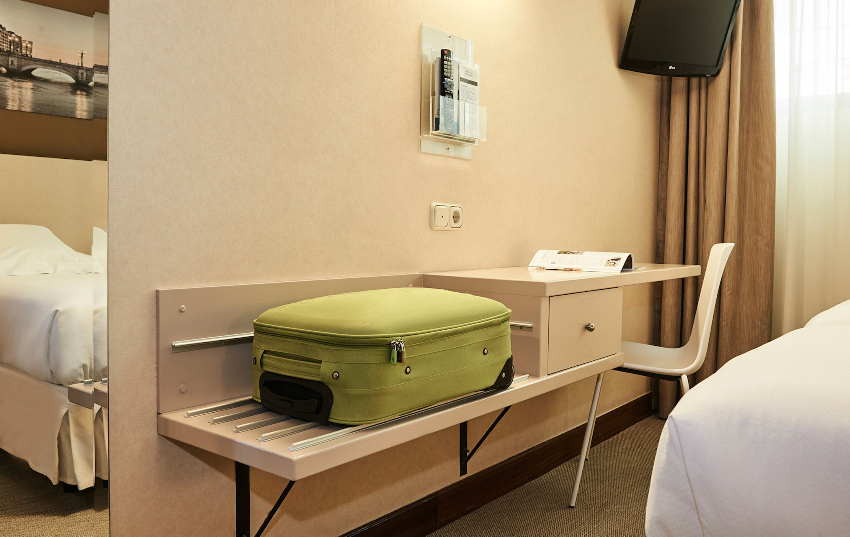 A hotel room with a bed, a desk with a green suitcase, a chair, a flat-screen TV, and a wall-mounted information board.