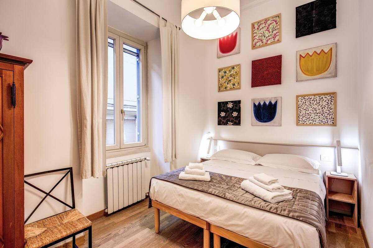 Un dormitorio ordenado con cama, mesitas de noche, una silla y una ventana con cortinas.