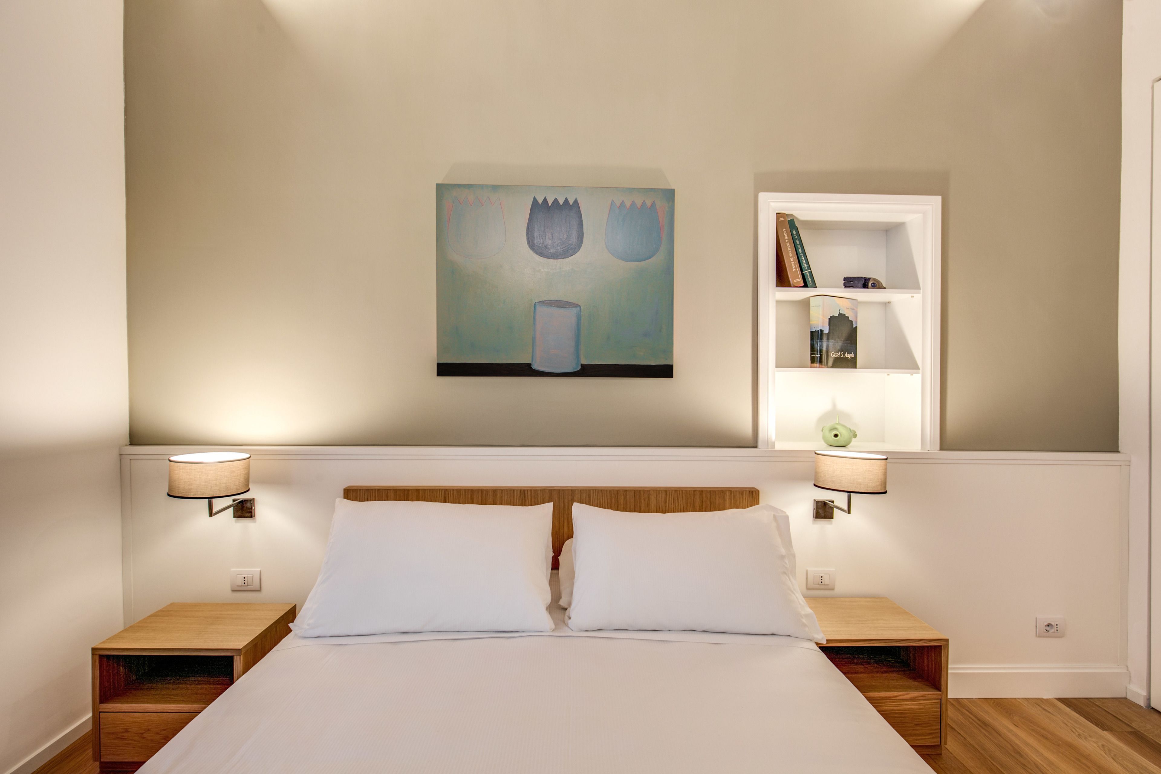 A neatly made bed with two pillows, a wall-mounted painting, a bookshelf with books and decorative items, and two bedside lamps.